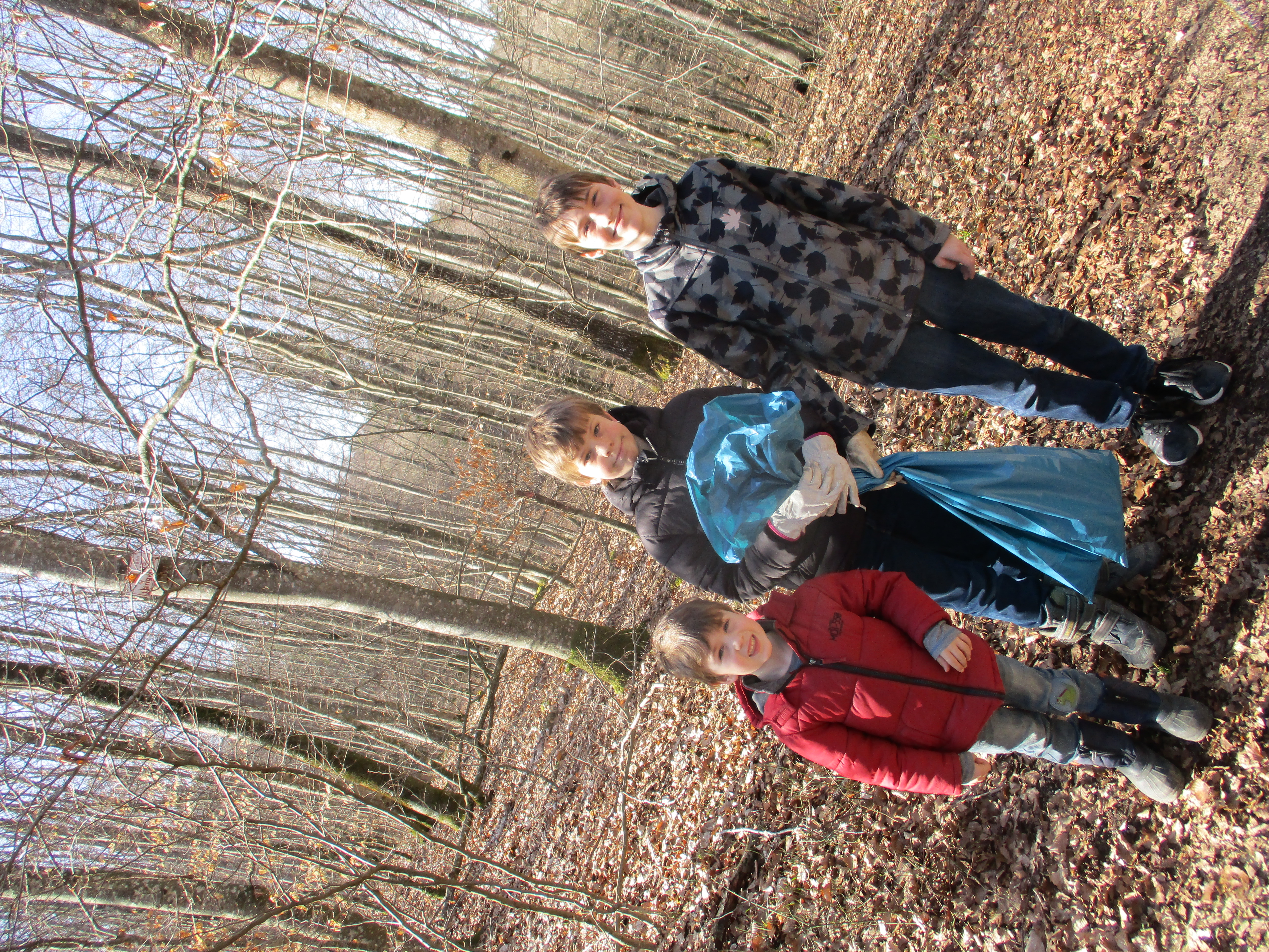 3 Kinder in einem Laubwald, die Bäume tragen kein Laub, die Sonne scheint, ein Kind trägt einen blauen Müllsack