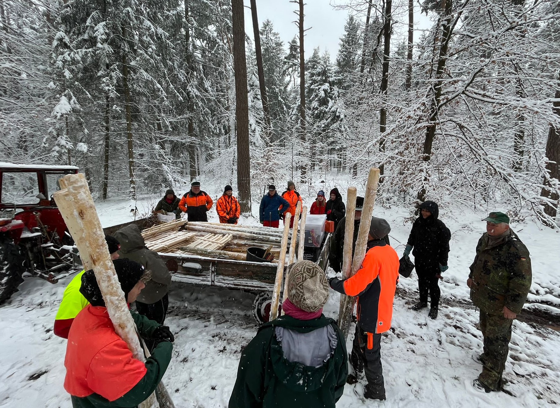 Eine Gruppe Menschen stehen im verschneiten Wald um einen Anhänger voller Baumaterial für Zäune