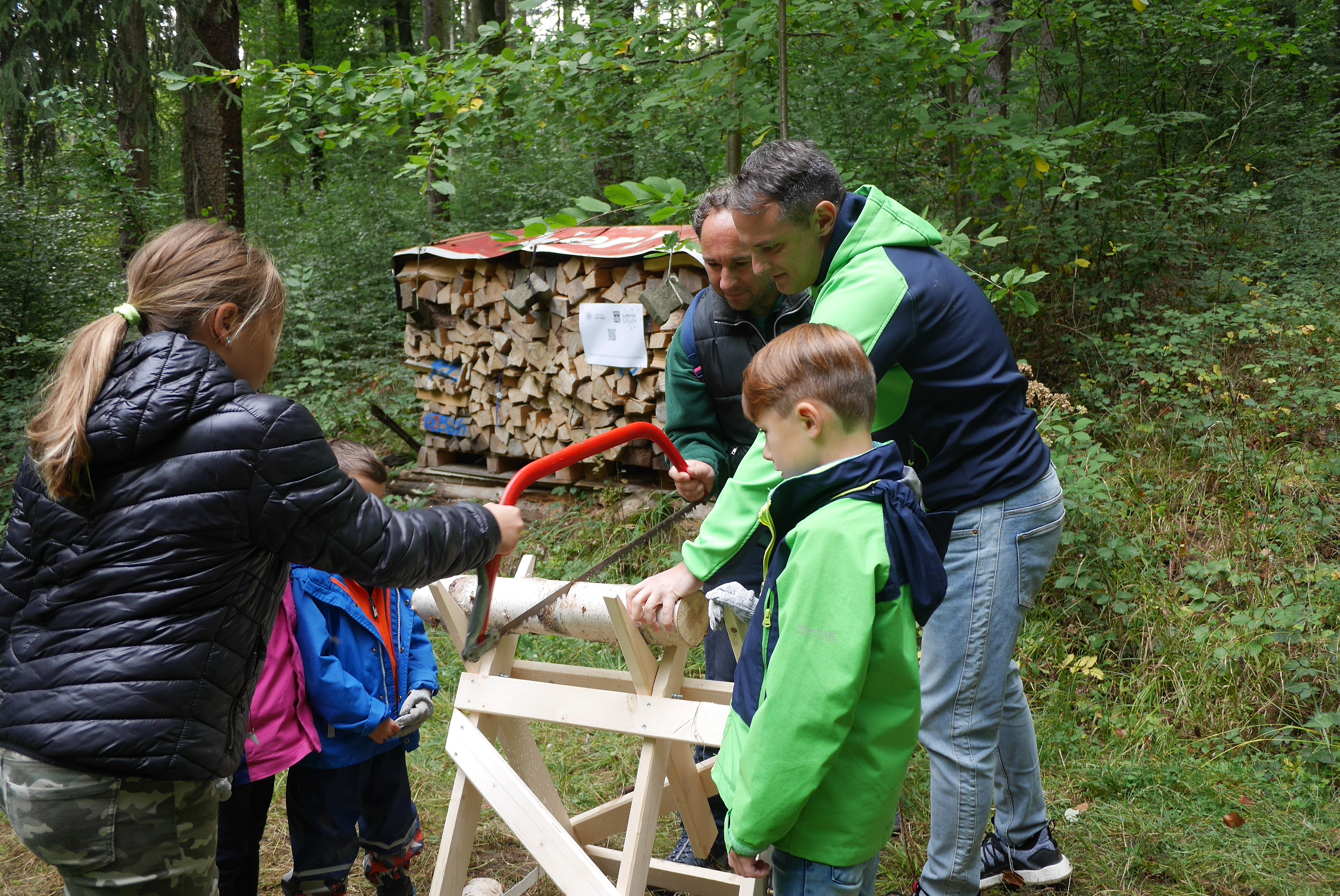 2 Erwachsene und mehrere Kinder stehen am Sägebock und Sögen einen Baumstamm. 